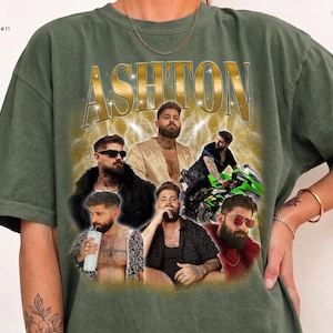 May include: A moss green t-shirt featuring the name ASHTON in gold lettering at the top. The graphic includes multiple photos of a bearded man in various outfits and poses, including one on a green motorcycle, set against a gold lightning background.