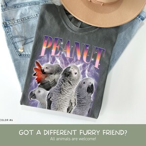 May include: A charcoal gray t-shirt featuring the word PEANUT in a gradient purple and orange font above three African Grey parrots. The design includes a purple lightning background. The shirt is displayed with blue jeans and a tan hat.