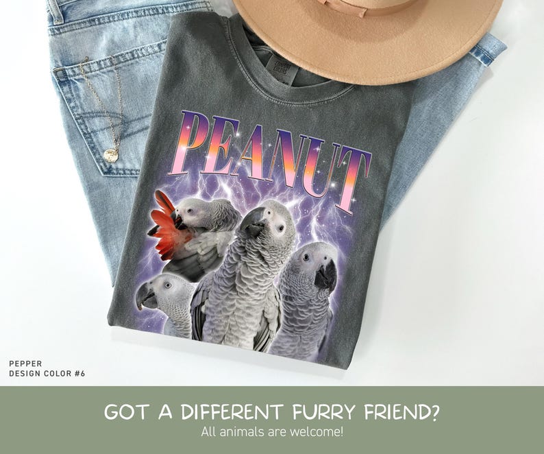 May include: A charcoal gray t-shirt featuring the word PEANUT in a gradient purple and orange font above three African Grey parrots. The design includes a purple lightning background. The shirt is displayed with blue jeans and a tan hat.