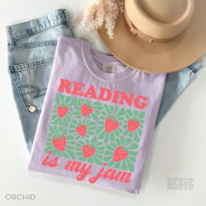 May include: A light purple t-shirt with the text "READING is my jam" in pink. The shirt features a pattern of strawberries and green leaves. The shirt is folded on top of a pair of blue jeans, a tan hat, and a necklace.