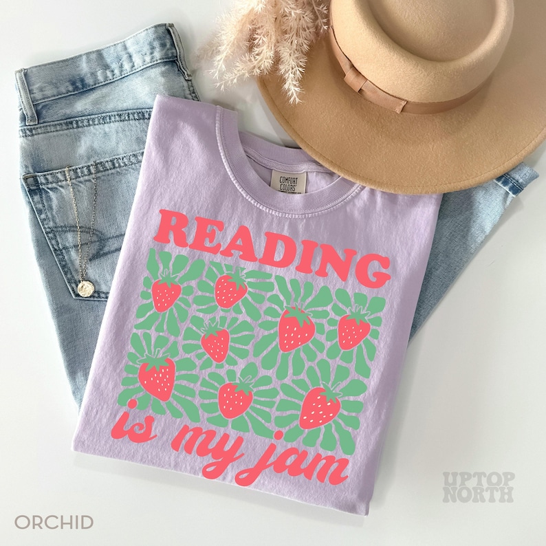 May include: A light purple t-shirt with the text "READING is my jam" in pink. The shirt features a pattern of strawberries and green leaves. The shirt is folded on top of a pair of blue jeans, a tan hat, and a necklace.