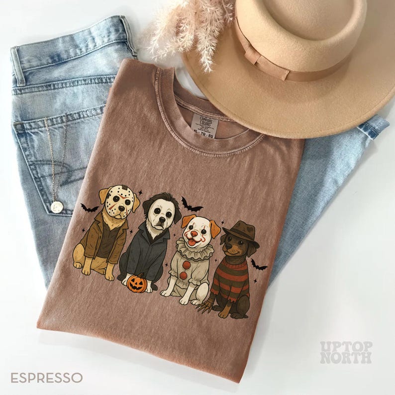 May include: Espresso-colored t-shirt featuring a Halloween-themed graphic of four dogs dressed as horror movie characters. The shirt is paired with light blue jeans, a necklace, and a tan hat. The text "UPTOP NORTH" is visible.