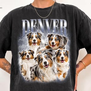 May include: A black short-sleeve t-shirt featuring the word DENVER in large silver metallic-style lettering at the top. Below the text is a graphic collage of five Australian Shepherd dogs with blue merle and brown fur, set against a hazy, glowing background.