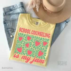 K&ouml;nnte beinhalten: Ein gelbes T-Shirt mit dem Text "SCHOOL COUNSELING is my jam" in Pink, zusammen mit Erdbeer-Grafiken. Das Shirt wird mit hellblauen Jeans und einem beigen Hut kombiniert. Das Wort "BANANA" ist unten links aufgedruckt.