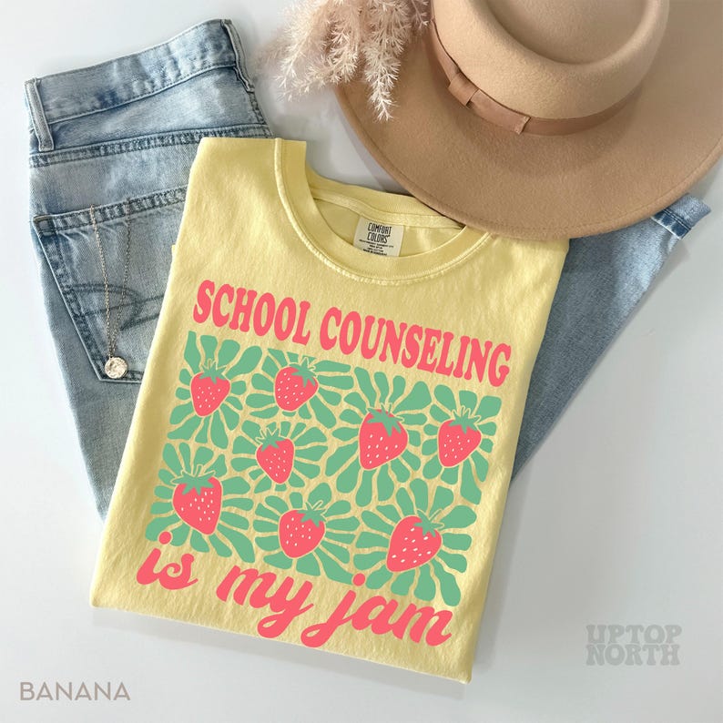 K&ouml;nnte beinhalten: Ein gelbes T-Shirt mit dem Text "SCHOOL COUNSELING is my jam" in Pink, zusammen mit Erdbeer-Grafiken. Das Shirt wird mit hellblauen Jeans und einem beigen Hut kombiniert. Das Wort "BANANA" ist unten links aufgedruckt.