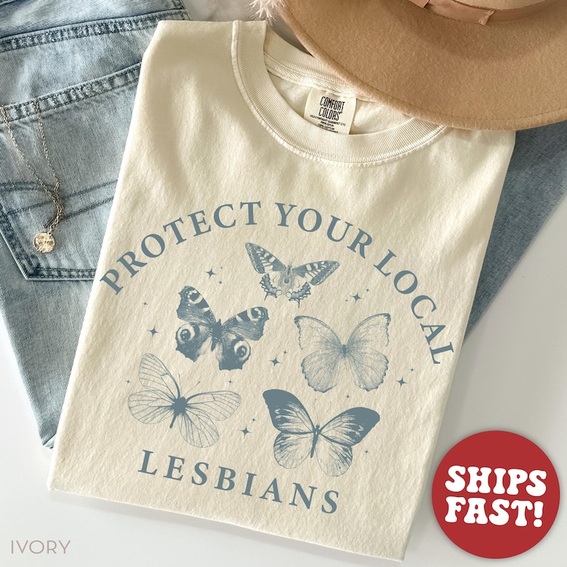 May include: Ivory-colored t-shirt with the text "PROTECT YOUR LOCAL LESBIANS" in blue, along with butterfly illustrations. The shirt is folded, with a hat and jeans in the background.