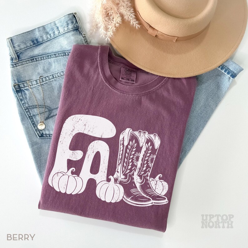May include: A berry-colored t-shirt with a white graphic that says "FALL" with pumpkins and cowboy boots. The shirt is laid on top of light-wash jeans, with a tan hat and a gold necklace. The word "BERRY" is printed at the bottom left.