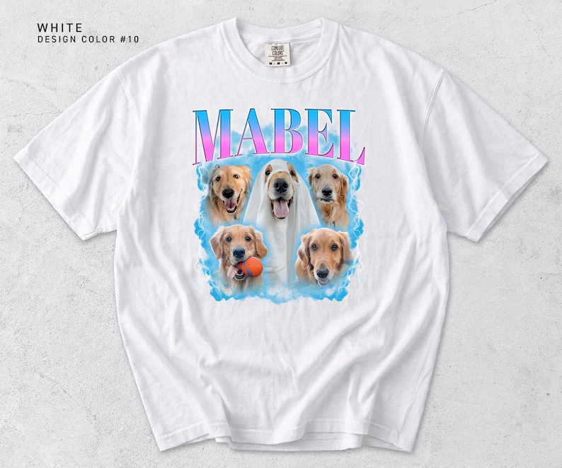 Pu&ograve; includere: T-shirt bianca con il nome "MABEL" in un font sfumato blu e rosa. La maglietta presenta una grafica di cinque cani golden retriever, alcuni con una palla, su uno sfondo di nuvole blu. Le parole "WHITE DESIGN COLOR #10" sono stampate in alto.