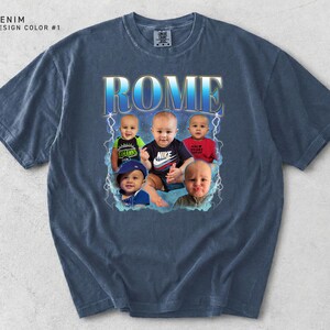 K&ouml;nnte beinhalten: Denimblaues T-Shirt mit einem grafischen Design mit dem Wort "ROME" in blauer und goldener Schrift, mit Bildern von f&uuml;nf Babys. Das Shirt hat einen Rundhalsausschnitt und kurze &Auml;rmel.