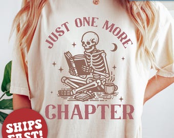 May include: A white t-shirt with a skeleton reading a book and a cup of coffee. The text on the shirt says "Just One More Chapter".