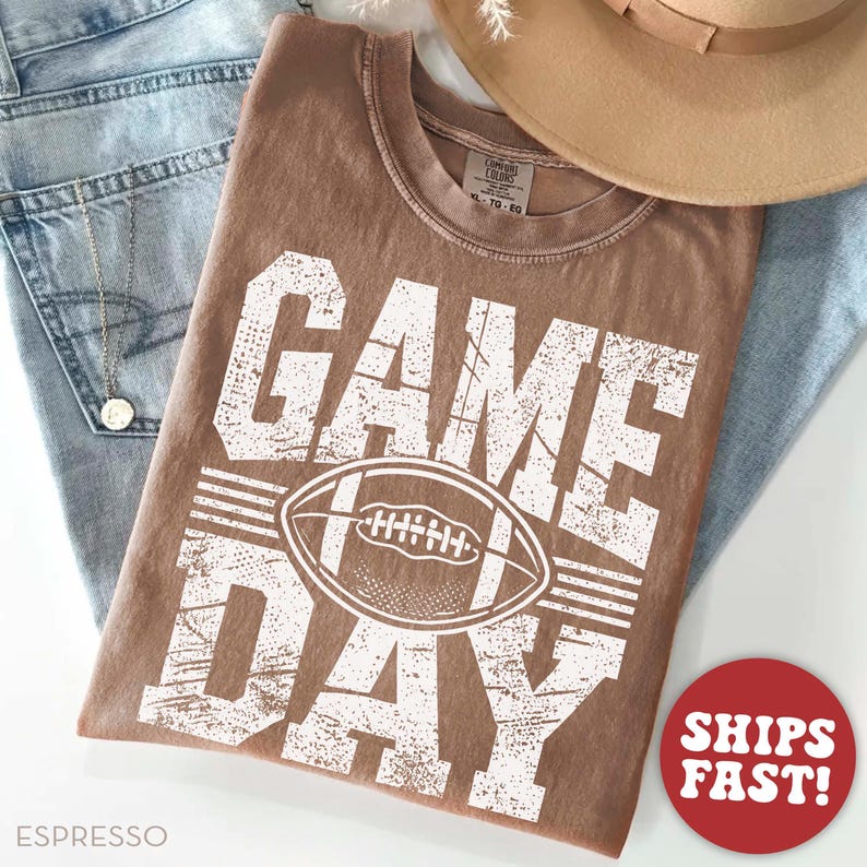 May include: Espresso-coloured t-shirt featuring the words "GAME DAY" and a white football graphic. The shirt is shown with blue jeans and a tan hat. The crew neck shirt has a distressed, vintage appearance.