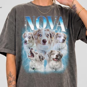 May include: A charcoal gray short-sleeve t-shirt featuring the word NOVA in large blue and white letters above a collage of five Australian Shepherd dogs. The graphic includes a blue glowing aura and small white star accents.