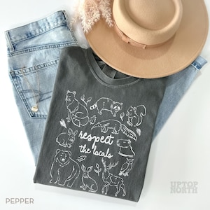 K&ouml;nnte beinhalten: Draufsicht auf ein Outfit mit einem dunkelgrauen T-Shirt mit wei&szlig;em Tierdruck und dem Text "respect the locals". Dazu eine hellblaue Jeans, ein beigefarbener Hut und eine Halskette. Das Wort "PEPPER" ist sichtbar. Der Look ist l&auml;ssig und naturverbunden.