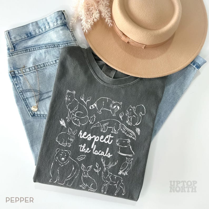 K&ouml;nnte beinhalten: Draufsicht auf ein Outfit mit einem dunkelgrauen T-Shirt mit wei&szlig;em Tierdruck und dem Text "respect the locals". Dazu eine hellblaue Jeans, ein beigefarbener Hut und eine Halskette. Das Wort "PEPPER" ist sichtbar. Der Look ist l&auml;ssig und naturverbunden.