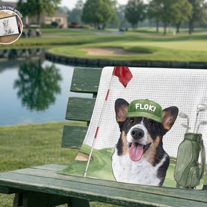 Op de afbeelding: Een witte handdoek met wafelstructuur met een kleurrijke illustratie van een zwart, wit en bruine Corgi die een groene honkbalpet draagt met de naam FLOKI erop, naast een golftas met clubs en een rode vlag op een golfbaan.