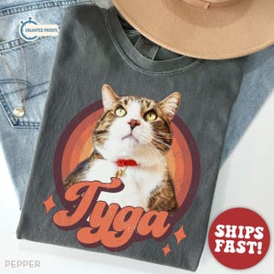May include: Dark gray t-shirt featuring a cat portrait inside a retro-style rainbow circle. The cat has brown and white fur, green eyes, and a red collar. The name "Tyga" is written in orange script below the cat.