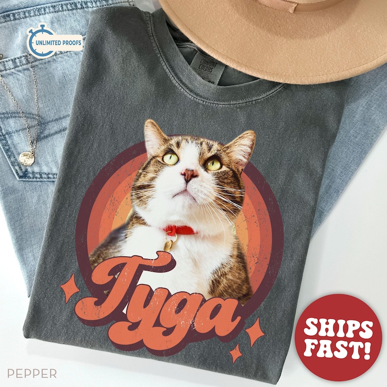 May include: Dark gray t-shirt featuring a cat portrait inside a retro-style rainbow circle. The cat has brown and white fur, green eyes, and a red collar. The name "Tyga" is written in orange script below the cat.