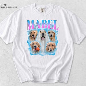 May include: A white short-sleeve t-shirt featuring a graphic print of five golden retriever dogs. The name MABEL is printed in large blue and pink gradient letters above the dogs. One dog wears a white veil, and another holds an orange ball.