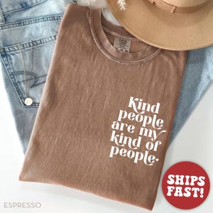May include: Espresso-colored t-shirt with the text "Kind people are my kind of people" in white. The shirt is folded on top of blue jeans and a tan hat. A red circle with "SHIPS FAST!" is in the bottom right corner.