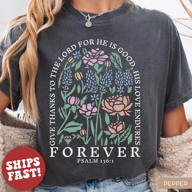 May include: Dark gray t-shirt with a floral design in pastel colors. The design includes the text "GIVE THANKS TO THE LORD FOR HE IS GOOD, HIS LOVE ENDURES FOREVER" and "PSALM 136:1". The shirt is labeled "PEPPER".