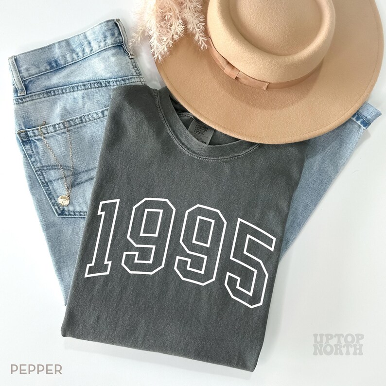 Puede incluir: Una camiseta gris oscura con el a&ntilde;o 1995 impreso en blanco en la parte delantera. La camiseta est&aacute; colocada sobre un fondo de mezclilla azul claro con un sombrero marr&oacute;n y una rama de flores secas.
