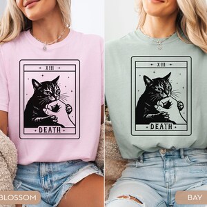 Op de afbeelding: Twee t-shirts, &eacute;&eacute;n roze en &eacute;&eacute;n groen, elk met een zwart-wit tarotkaartontwerp. De kaart toont een kat en een hand, met het woord "DEATH" onderaan. De bovenkant van de kaart leest "+ XIII +".