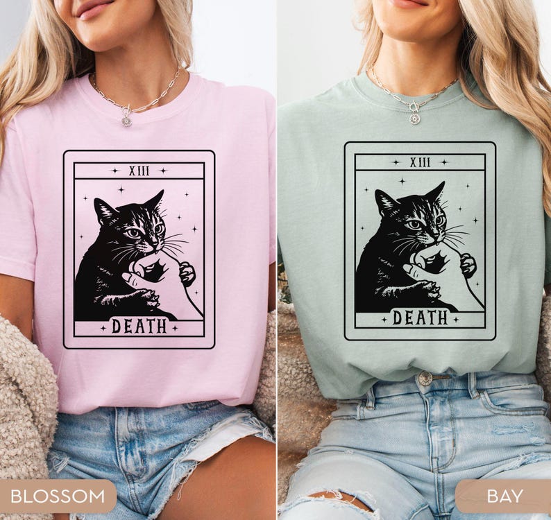 Op de afbeelding: Twee t-shirts, &eacute;&eacute;n roze en &eacute;&eacute;n groen, elk met een zwart-wit tarotkaartontwerp. De kaart toont een kat en een hand, met het woord "DEATH" onderaan. De bovenkant van de kaart leest "+ XIII +".