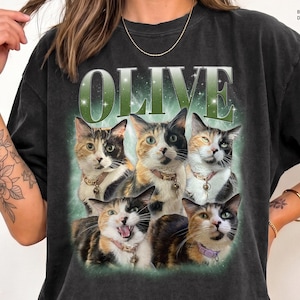May include: A black t-shirt featuring the word OLIVE in large green letters above a graphic of five calico cats. The cats are shown in various poses, wearing collars with small charms, set against a soft green glowing background.
