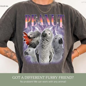 May include: A charcoal gray short-sleeve t-shirt featuring a graphic print of four African Grey parrots. The word PEANUT is printed in large, gradient-colored letters above the birds, set against a purple background with lightning bolt accents.