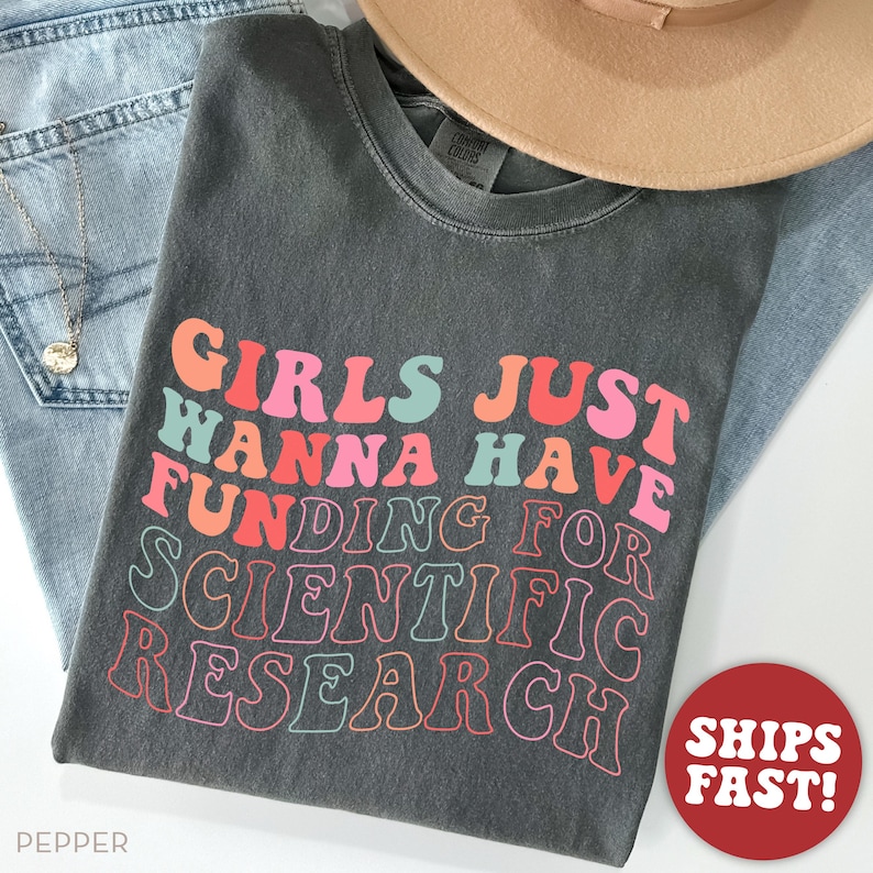 Pode incluir: Uma camiseta cinza escura com o texto "Girls Just Wanna Have Fun-ding for Scientific Research" em cores do arco-&iacute;ris. O texto est&aacute; em um estilo de fonte retr&ocirc;.