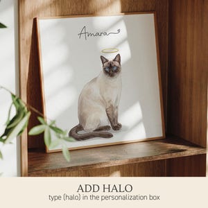 Op de afbeelding: Een vierkante ingelijste print met een aquarelillustratie van een Siamees met blauwe ogen en een gouden halo boven het hoofd. De naam Amara staat in zwarte cursieve letters boven de kat, vergezeld van een klein hartje.