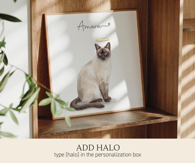 Op de afbeelding: Een vierkante ingelijste print met een aquarelillustratie van een Siamees met blauwe ogen en een gouden halo boven het hoofd. De naam Amara staat in zwarte cursieve letters boven de kat, vergezeld van een klein hartje.