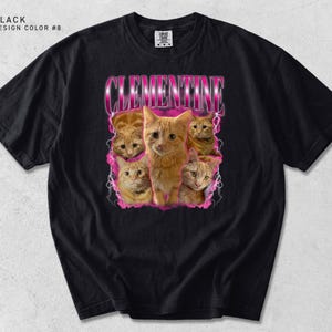 Pu&ograve; includere: T-shirt nero con una grafica di gatti tabby arancioni e il nome "CLEMENTINE" in un font rosa retr&ograve;. La maglietta ha un girocollo e maniche corte. Il testo "BLACK DESIGN COLOR #8" &egrave; in alto a sinistra.