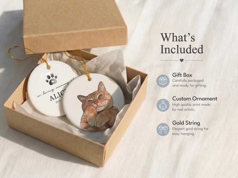 May include: Two round white ceramic ornaments with gold strings inside a brown cardboard gift box. One ornament features a brown tabby cat illustration, while the other displays a black paw print and the text in loving memory ALICE 2010.
