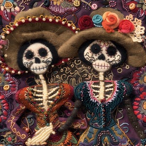 May include: Two felt skeleton figures wearing traditional Mexican clothing and sombreros. The figures are decorated with beads and embroidery. The background is a purple fabric with floral embroidery.