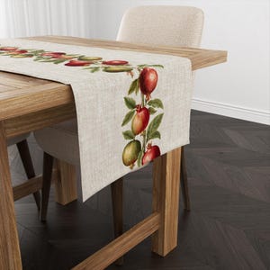 May include: A beige linen table runner with a repeating pattern of green leaves and red pomegranates. The runner is on a wooden table with a chair.