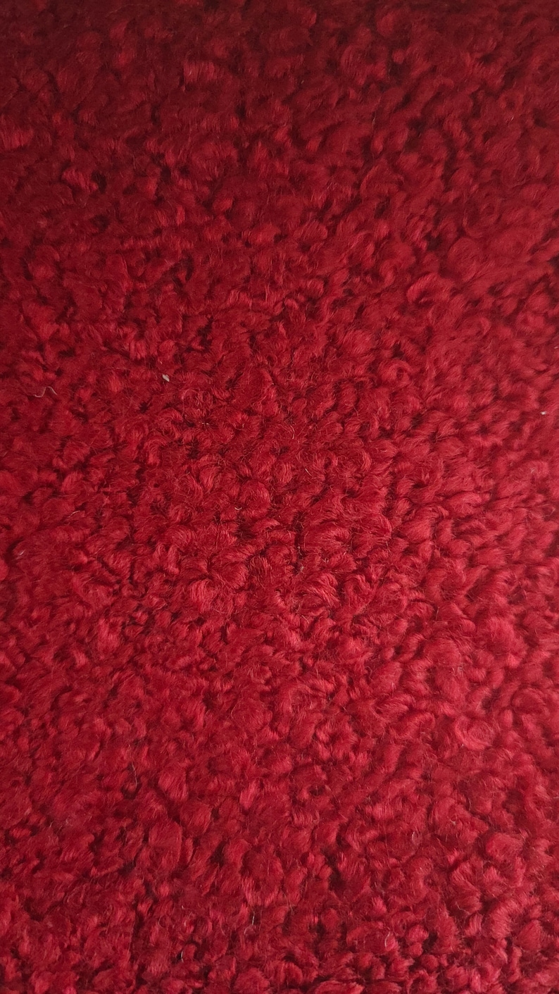 May include: Close-up of a textured, deep red fabric. The material has a dense, looped pile, creating a plush, uneven surface. The color is a rich, saturated shade of red, with subtle variations in tone across the surface.
