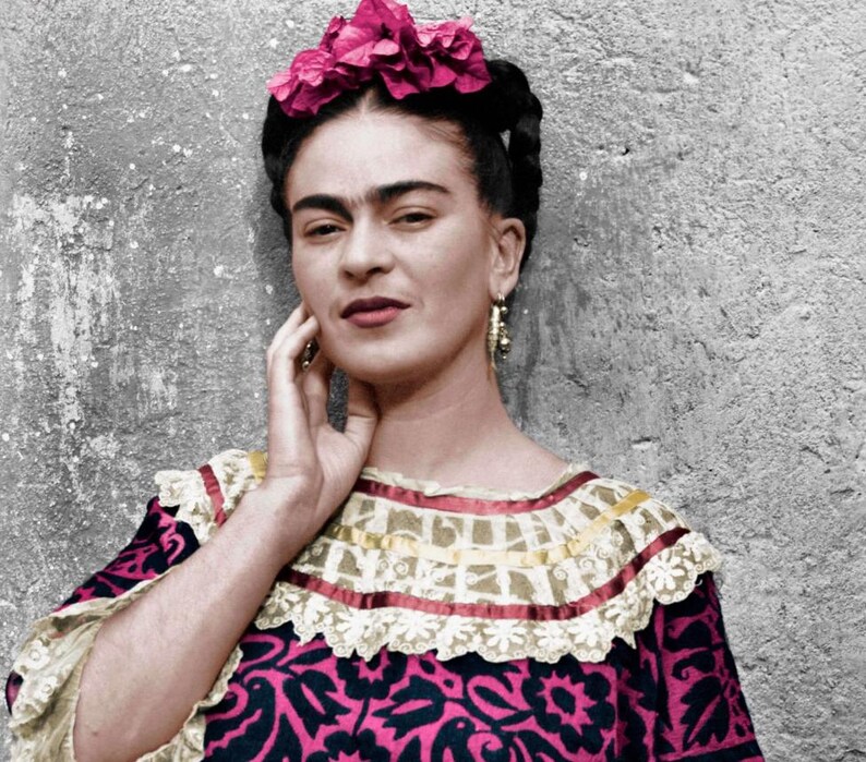 May include: A portrait of a person with dark hair, wearing a pink floral headpiece and gold earrings. The person is wearing a patterned dress with a ruffled collar. The background is a textured gray wall.