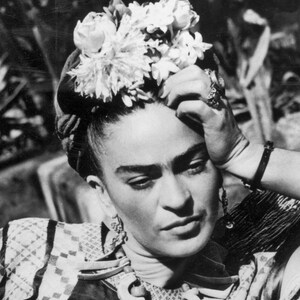May include: Black and white portrait of a person with dark hair, a floral headpiece, and ornate jewelry. The person is wearing a patterned top and has their hand resting on their forehead, with a contemplative expression.