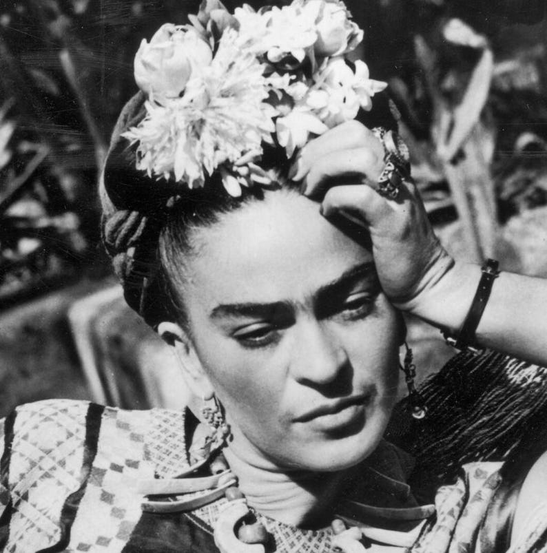 May include: Black and white portrait of a person with dark hair, a floral headpiece, and ornate jewelry. The person is wearing a patterned top and has their hand resting on their forehead, with a contemplative expression.