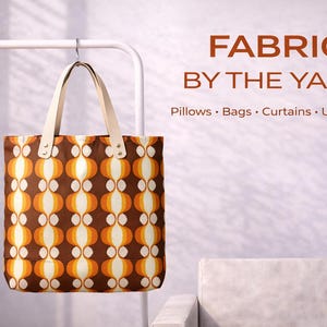May include: A tote bag with a retro pattern of brown, orange, and cream-coloured shapes hangs from a white metal rack. The bag has cream-coloured handles. Text on the right reads "FABRIC BY THE YARD" and lists items like pillows and bags.