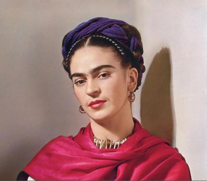 May include: A portrait of a person wearing a purple head wrap, a red shawl, and gold hoop earrings. The person is also wearing a necklace with a white and gold design. The background is a neutral color.
