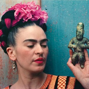 May include: A portrait of a person with dark hair styled with braids and adorned with a pink flower crown. They are holding a small, green-toned statue of a seated figure. The person is wearing a colorful embroidered top and gold earrings.