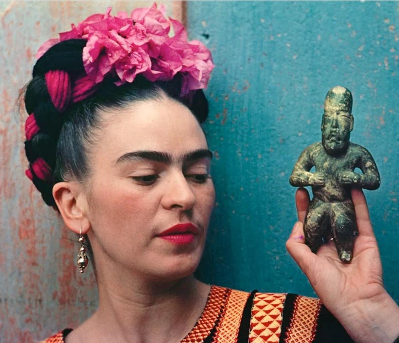 May include: A portrait of a person with dark hair styled with braids and adorned with a pink flower crown. They are holding a small, green-toned statue of a seated figure. The person is wearing a colorful embroidered top and gold earrings.