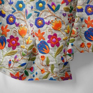 Pode incluir: Close-up de um tecido com bordado floral colorido. O design apresenta flores azuis, laranja e rosa com folhas e caules verdes sobre um fundo cinza claro. O bordado é detalhado e vibrante.