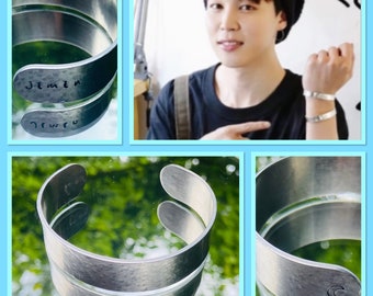 May include: A silver metal cuff bracelet with the word "Jimin" engraved on one side and a crescent moon on the other side.