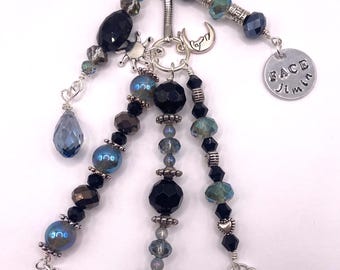 May include: A silver keychain with multiple beaded strands in shades of blue, black, and silver. Charms include a blue butterfly, a sun, a crescent moon, a star, and two round charms with text and a hand gesture. The beads are various shapes and sizes.
