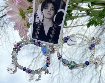 May include: Two beaded bracelets with silver charms. One bracelet has a camera charm, a crescent moon charm, a star charm, and a charm with two hands clasped together. The other bracelet has a heart charm with the word "filter" written on it, a bird charm, and a charm with a circle and a line through it. The bracelets are on a white surface with a blurred image of a person in the background.