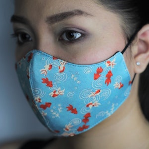 May include: A blue face mask with a pattern of red and white fish swimming in circles. The mask is worn by a person with dark hair.
