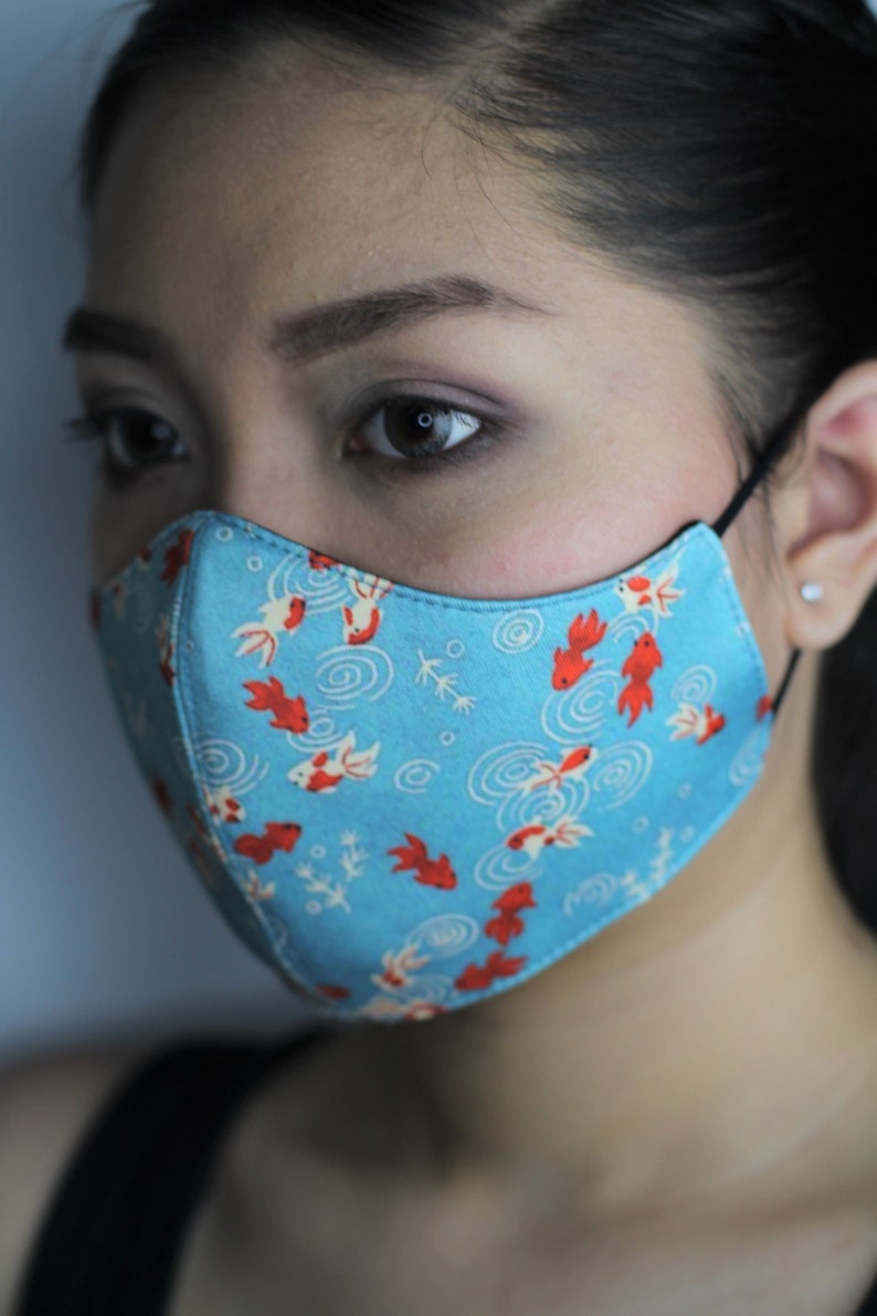May include: A blue face mask with a pattern of red and white fish swimming in circles. The mask is worn by a person with dark hair.